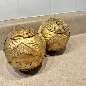 May include: Two gold-colored decorative spheres with leaf and spiral rope designs. Each sphere has a loop for hanging. The spheres have a distressed, antique appearance, suggesting they are vintage or handcrafted.