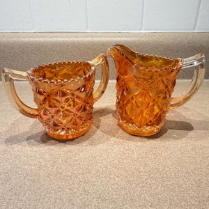 May include: Two antique orange glass cream and sugar set. The pieces have a diamond pattern and clear handles. The rims are scalloped. The set is displayed on a neutral surface.