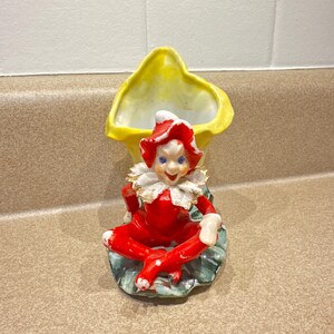May include: A vintage ceramic figurine of a seated elf in a red outfit with a white collar and hat. The elf is positioned in front of a yellow, flower-shaped vase. The figurine has blue eyes and is resting on a green base.