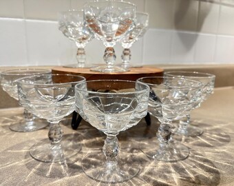 May include: A collection of clear, cut-glass stemmed dessert or cocktail glasses. The glasses have a diamond-cut pattern on the bowl and a decorative stem. Some glasses are arranged on a small wooden stand.