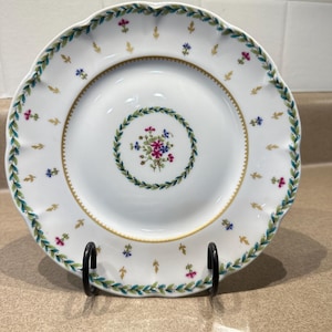 May include: White ceramic plate with a scalloped edge, featuring a floral and leaf design in green, blue, pink, and gold. The plate has a central floral arrangement and is displayed on a black metal stand.