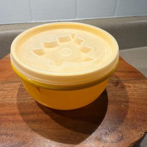 May include: A yellow plastic bowl with a matching lid. The lid has a decorative design in the center. The bowl is sitting on a wooden surface.