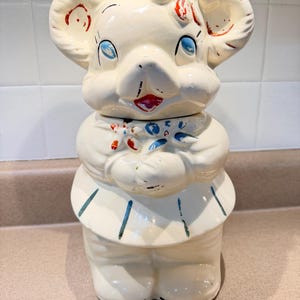May include: Cream-colored ceramic cookie jar in the shape of a cartoon elephant. The elephant has large ears, blue eyes, and red and blue accents. The jar is in the form of a standing elephant with a skirt and arms crossed.