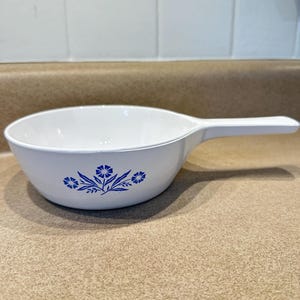 May include: White ceramic serving bowl with a handle, featuring a blue floral design. The bowl has a rounded shape and a long, integrated handle. The floral pattern includes stylized flowers and leaves.