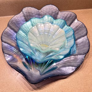May include: A set of three decorative glass bowls in varying shades of blue and iridescent hues. The bowls are nested, with a large, scalloped-edged bowl at the base, followed by two smaller bowls. The glass has a rippled texture.