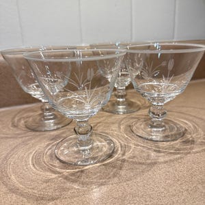May include: Set of four clear, stemmed cocktail glasses. Each glass features a delicate etched floral design around the bowl and a rounded stem. The glasses are arranged on a light brown countertop, with a white tiled backsplash.