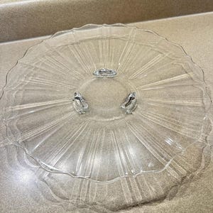 May include: A clear glass cake stand with a scalloped edge and a starburst design radiating from the center. The stand has three small feet and a handle. The stand is made of clear glass.