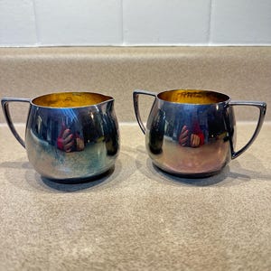 May include: Two silver-colored cream pitchers with a shiny, reflective surface. Each pitcher has a rounded body, a small handle, and a gold-colored interior. The pitchers are placed side by side on a neutral-colored surface.