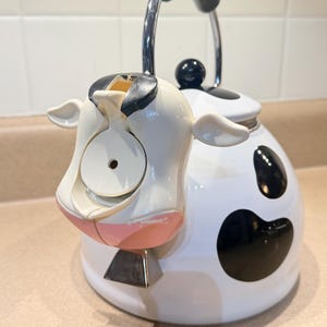 May include: A whimsical white and black cow-shaped tea kettle with a black handle and spout. The kettle features a cow face design with pink accents and black spots. The handle is made of black plastic and chrome. The kettle is designed to sit on a stovetop.