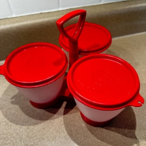 May include: A red and white plastic condiment caddy with three round containers. Each container has a red lid and a white base. The caddy has a red handle for easy carrying. The containers are designed for holding various condiments.