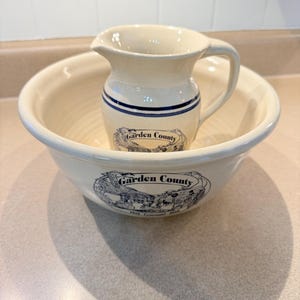 May include: Cream-colored ceramic pitcher and bowl set. The pitcher has a handle and a spout, with blue stripes and a "Garden County" emblem. The bowl is wide and shallow, also with the "Garden County" emblem. A vintage-style kitchenware set.