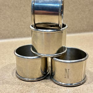 May include: Four silver napkin rings stacked on a neutral surface. Three rings are visible, with one stacked on top. One ring is engraved with the letter "N". The rings have a simple, elegant design.