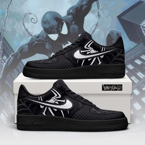 May include: A pair of black sneakers featuring a white Nike swoosh, a white spider graphic, and a black spiderweb pattern on the side panels. The shoes have black laces, a black sole, and are displayed on a white box with the text Luxe's Fun.