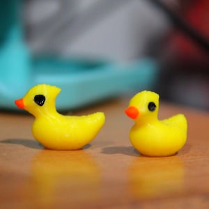 May include: Two small, yellow duck figurines with orange beaks and black eyes. The ducks are on a wooden surface. One duck is slightly larger than the other. The background is blurred, with hints of blue and red.