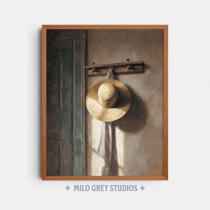 Op de afbeelding: Een ingelijste prent toont een strohoed die aan een wandhaak hangt. De beige hoed hangt aan een lichtgekleurde stof. De muur is neutraal van kleur en links is een deur zichtbaar. De prent is van Milo Grey Studios.