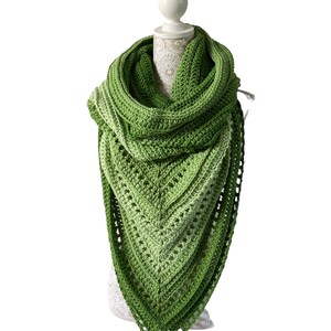 May include: A handmade, green crochet shawl displayed on a mannequin. The shawl features a triangular shape with a gradient of green hues, from light to dark. The open weave design adds texture and visual interest to the accessory.