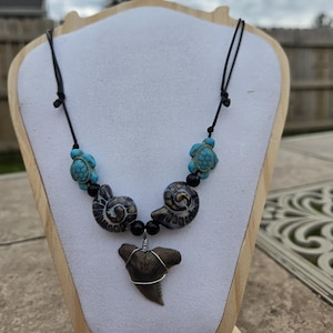 May include: A necklace featuring a brown shark tooth pendant wrapped in silver-colored wire, flanked by two dark spiral ammonite fossils, two bright turquoise sea turtle beads, and small black round beads on a black cord with adjustable knots.