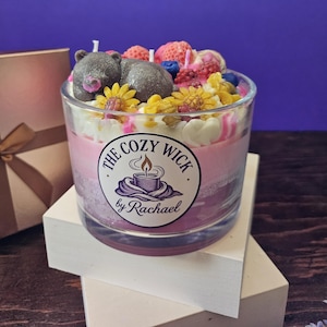 May include: A layered candle in a clear glass, featuring a decorative top with a gray bear, yellow flowers, and berries. The candle has pink and purple layers, with a label that reads "The Cozy Wick by Rachael."