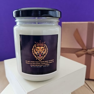 Puede incluir: Una vela blanca en un tarro de cristal con tapa negra. La etiqueta presenta una cabeza de león con corona y texto de Mateo 10:34. La vela está sobre un bloque blanco con fondo morado.