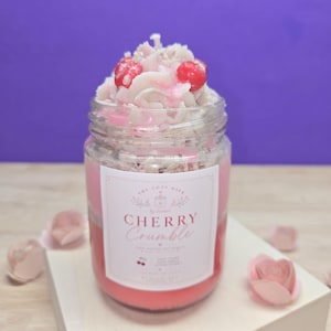May include: A jar candle with a cherry crumble design. The candle has a pink and white layered appearance, topped with decorative frosting and red cherry accents. The label reads "Cherry Crumble".