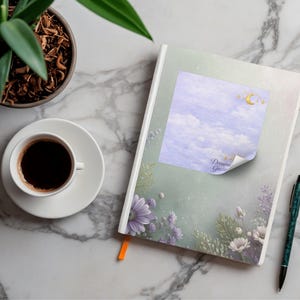 May include: A journal with a watercolor design featuring a sky scene with a moon and stars, and the text "Dream Guide". A cup of coffee, a pen, and a potted plant are also on the marble surface.