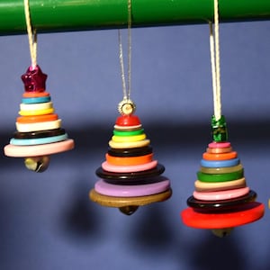 May include: Five handmade Christmas ornaments shaped like trees, crafted from colorful buttons. Each ornament has a bell at the bottom and is suspended by a string. The ornaments are in various colors, including pink, green, and yellow.
