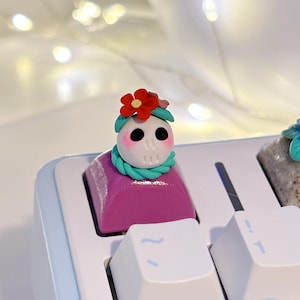 Puede incluir: Una tecla de teclado personalizada con un diseño de calavera caprichoso. La calavera tiene una corona de flores rojas y una bufanda turquesa, sobre una base magenta. La tecla forma parte de un teclado azul claro y blanco.