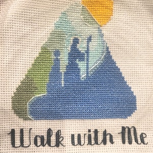 Può includere: Ricamo a punto croce con una silhouette di figure contro uno sfondo montuoso, con un sole giallo. Il disegno è nei toni del blu, verde e bianco, con le parole "Walk with Me" ricamate in nero sotto.