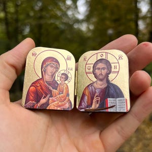 May include: Two small, gold-colored religious icons are held in a hand. The icons depict religious figures with detailed artwork. The icons have hinged lids and are rectangular in shape. The background is blurred, with green and brown tones.