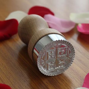 May include: A wooden stamp with a decorative, clear, patterned base and a silver band. The stamp is surrounded by red, pink, and white rose petals, arranged on a wooden surface.