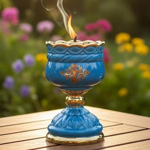May include: A lit blue ceramic oil lamp with gold accents sits on a wooden table. The lamp has a decorative floral design and is emitting smoke. The background features a blurred garden with colorful flowers and sunlight.