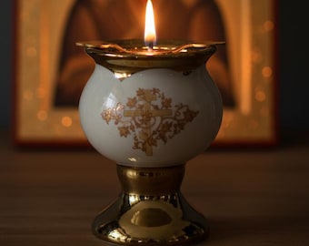 May include: A lit candle in a white and gold ceramic holder with a floral design. The candle holder is in front of a religious icon with a blurred background. The candle flame is bright.