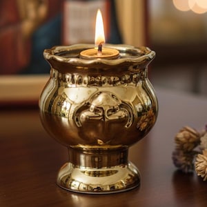 Op de afbeelding: Een goudkleurige kandelaar met een brandende kaars, geplaatst op een donker houten oppervlak. De kandelaar heeft een decoratief ontwerp. Op de achtergrond is een religieuze icoon te zien.