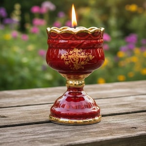 Op de afbeelding: Een brandende rode kaars in een decoratieve houder met gouden rand. De kaarsenhouder heeft een voetstuk en sierlijke gouden details. De vlam is helder tegen een vage achtergrond van groene en paarse bloemen.
