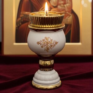 May include: A lit candle in a gold-rimmed holder sits in front of a religious icon. The candle holder is white with gold accents and a floral design. The icon depicts a religious figure in red and gold.