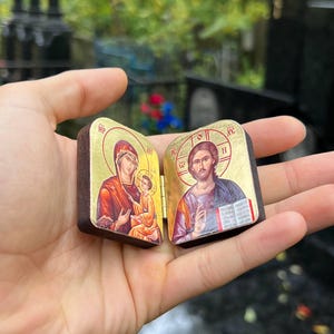 May include: A small, hinged wooden icon featuring religious imagery. The left panel depicts the Virgin Mary and child, while the right panel shows Jesus. Gold accents frame the images, and the wood has a dark brown finish.