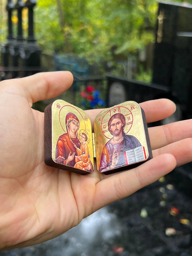 May include: A small, hinged wooden icon featuring religious imagery. The left panel depicts the Virgin Mary and child, while the right panel shows Jesus. Gold accents frame the images, and the wood has a dark brown finish.