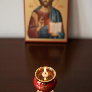 May include: A lit candle in a red and gold candle holder with a gold base. The candle holder is in front of a religious icon depicting a figure with a halo, holding a book. The background is blurred.