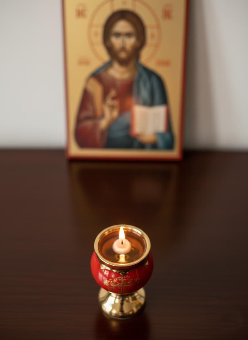 May include: A lit candle in a red and gold candle holder with a gold base. The candle holder is in front of a religious icon depicting a figure with a halo, holding a book. The background is blurred.