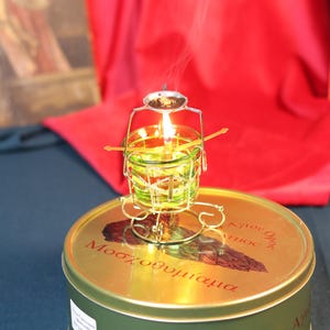 May include: A lit candle in a small, green glass container with a metal frame, sitting atop a round, gold-colored tin. The tin has red text and a brown illustration. Smoke rises from the candle against a red backdrop.
