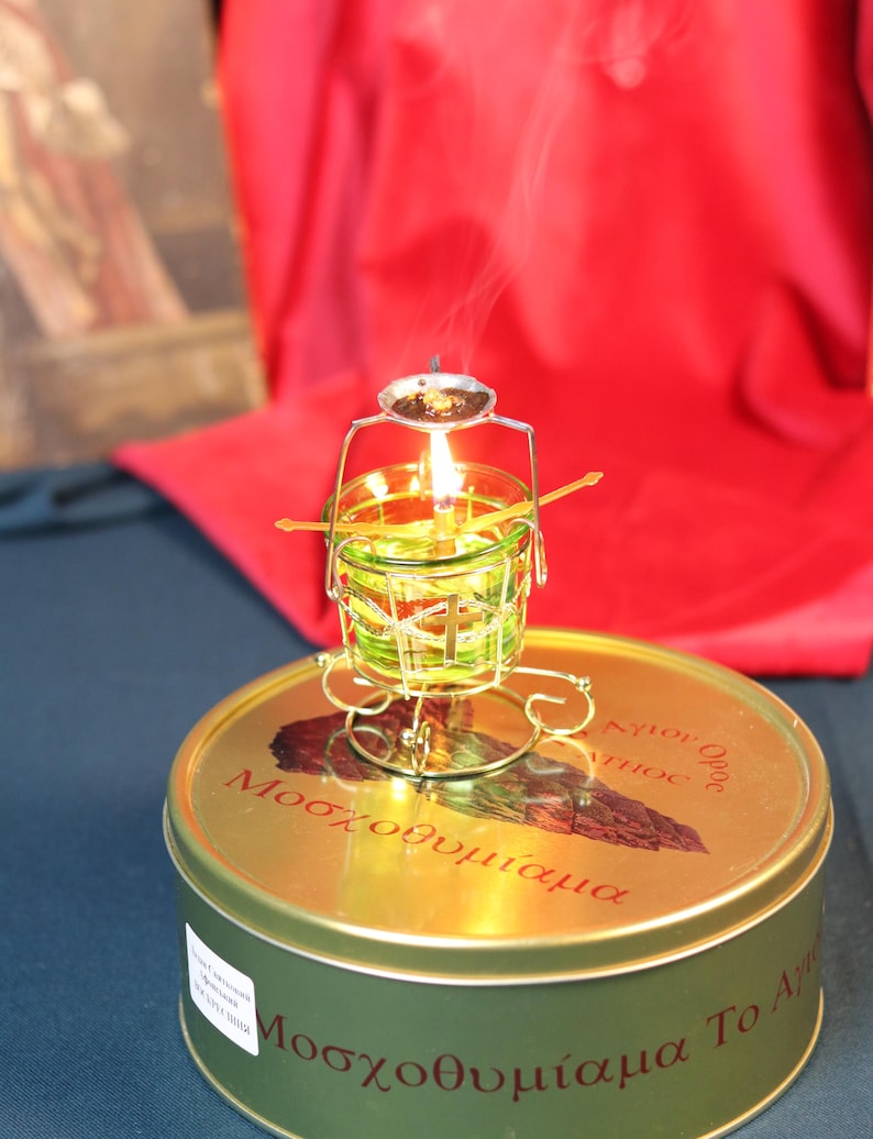 May include: A lit candle in a small, green glass container with a metal frame, sitting atop a round, gold-colored tin. The tin has red text and a brown illustration. Smoke rises from the candle against a red backdrop.