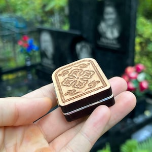May include: A small, square wooden box with a floral design carved into the light-colored lid. The box has dark brown sides and a silver band around the middle. The box is held in a person's hand.
