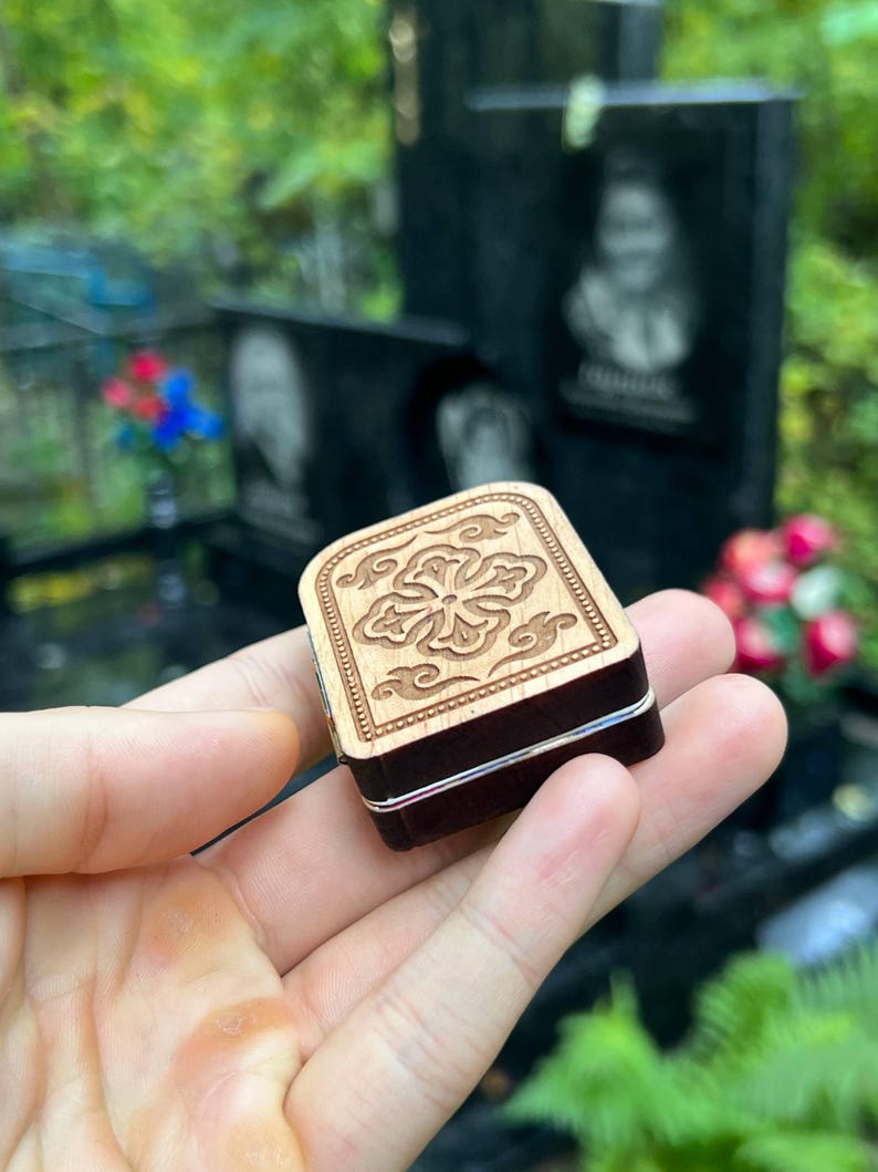 May include: A small, square wooden box with a floral design carved into the light-colored lid. The box has dark brown sides and a silver band around the middle. The box is held in a person's hand.