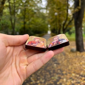 May include: Two small, rectangular religious icons are held in a hand. The icons feature detailed illustrations with gold backgrounds and dark brown frames. One depicts a figure holding a baby, and the other shows a robed figure.