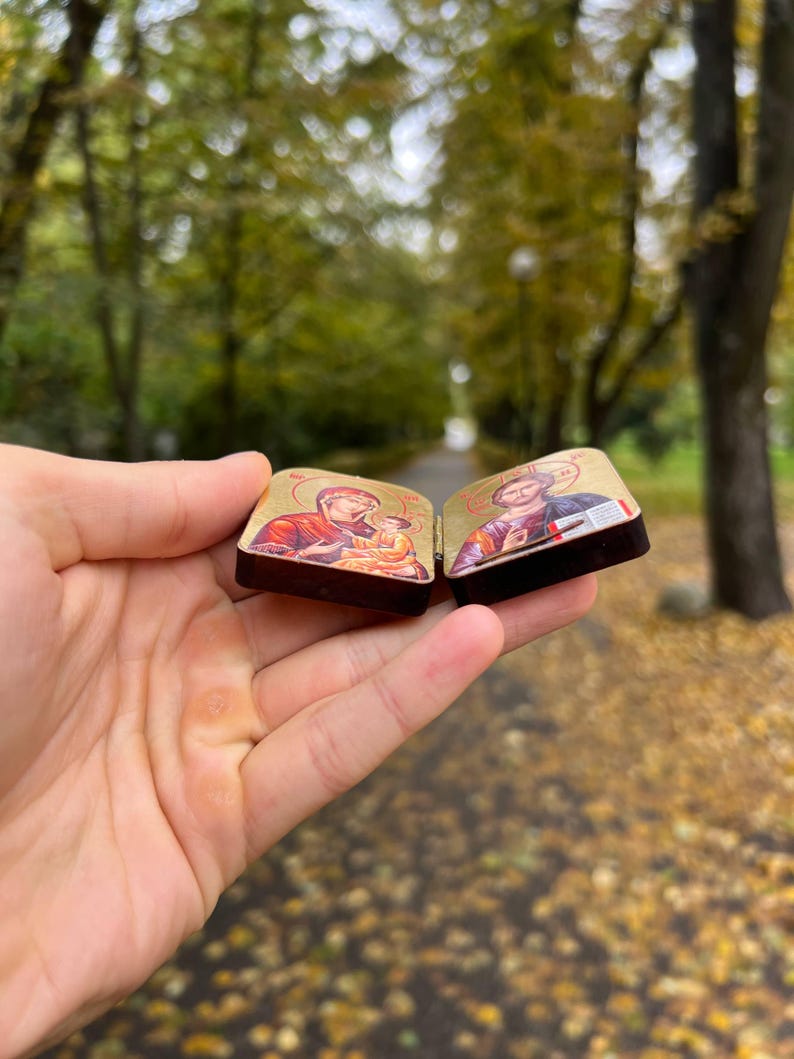 May include: Two small, rectangular religious icons are held in a hand. The icons feature detailed illustrations with gold backgrounds and dark brown frames. One depicts a figure holding a baby, and the other shows a robed figure.