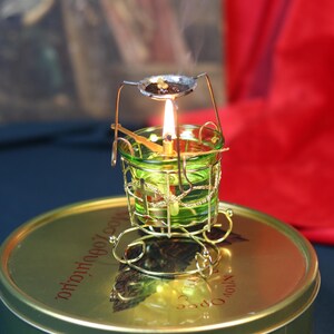 May include: A small, ornate incense burner with a lit candle inside a green glass holder. The holder is encased in a gold-colored wire frame, and a small metal dish above holds incense. The burner sits on a round, green tin.