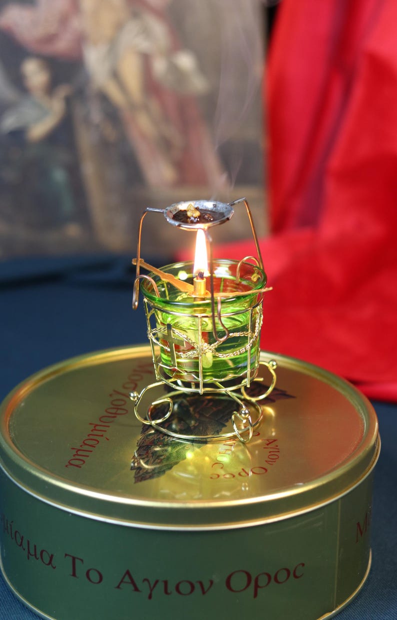 May include: A small, ornate incense burner with a lit candle inside a green glass holder. The holder is encased in a gold-colored wire frame, and a small metal dish above holds incense. The burner sits on a round, green tin.