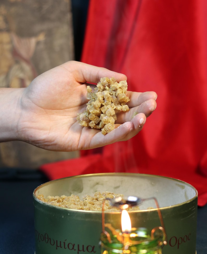 May include: A hand holding a cluster of small, light brown, granular pieces, possibly incense or a similar substance. The pieces are being sprinkled over a container. A small, lit candle is in the foreground, and a red cloth is in the background.
