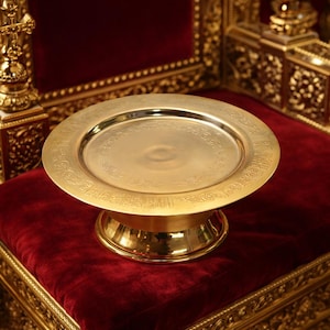 May include: A gold-coloured, round, decorative plate with a raised base. The plate has an ornate, engraved border and sits on a red velvet cushion. The background features gold-coloured architectural details.