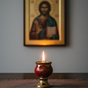 May include: A lit red and gold candle holder with a flame, set against a blurred background of a religious icon. The candle holder has a detailed design and sits on a wooden surface.